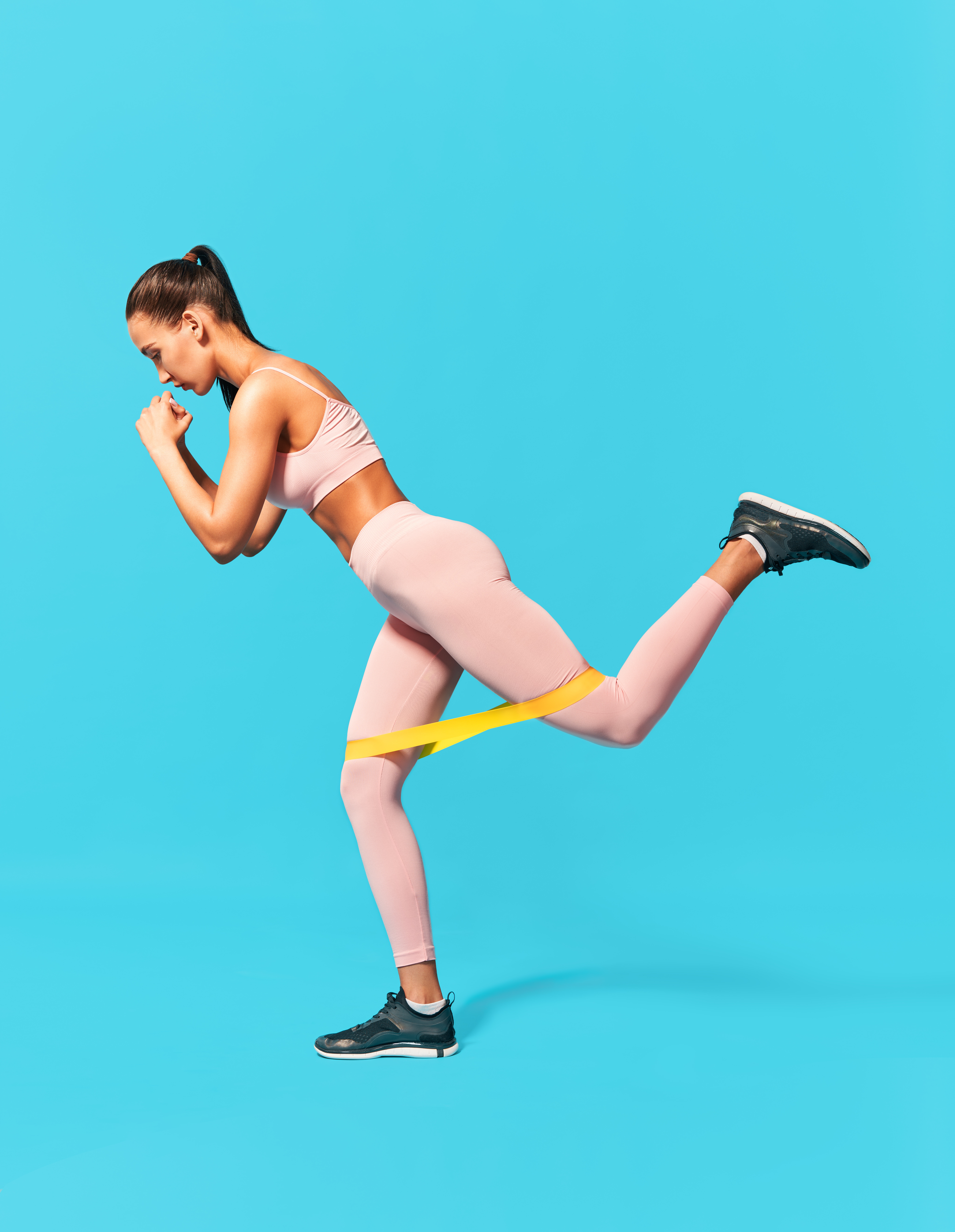 Frau in rosa Sportkleidung führt eine Kickback-Übung mit einem gelben Loop-Fitnessband vor türkisfarbenem Hintergrund aus.