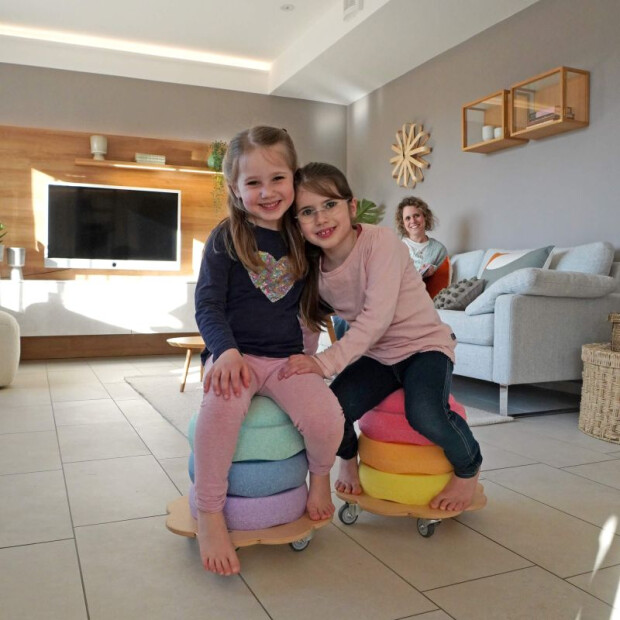 Anwendungsbild 3: Zwei Kinder sitzen gemeinsam auf Stapelstein-Türmen, die auf Pedalo® Rollflitzern im Wohnzimmer platziert sind.