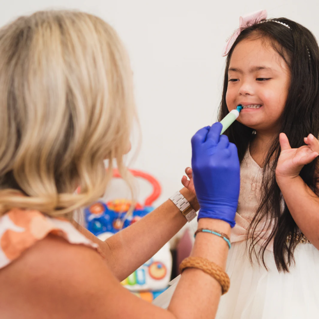 Kind sitzt frontal, schaut nach rechts, während eine erwachsene Person mit Handschuh ein Talktool-Gerät im Mundbereich anwendet, in Anwendungssituation vor hellem Hintergrund.